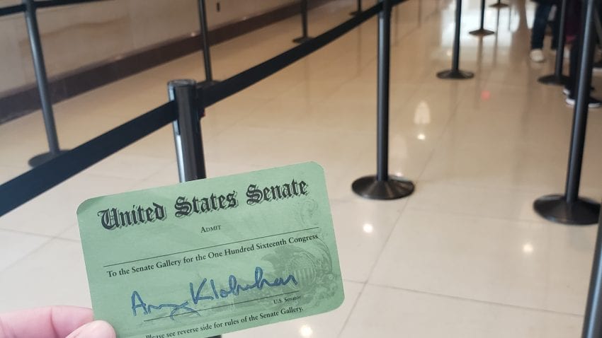 A picture of a green ticket for the Senate gallery in front of a tan floor with black ropes to separate the lines.
