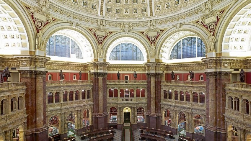 The Library of Congress Reading room, with several large half-circle windows and the top, and dozens of tables on the main floor. Books line the walls of the circular room.