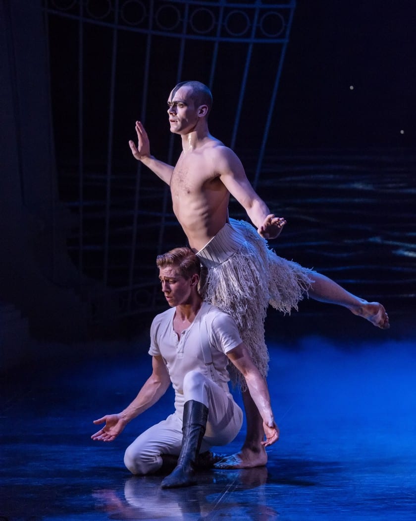 A dancer stands behind another performer sitting in front of him. The dancer in the front holds his arms open, while the standing dancer holds a leg up towards the back.