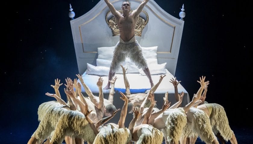 A male dancer stands under a spotlight on a white bed while other performers crouch around the bed with one arm up.