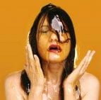 A photo of the artist covered in honey. The photo has an orange background and the artist's hands are up by her face. 