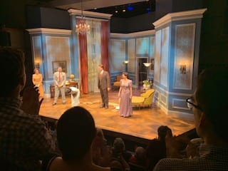 The stage of "Sheltered," featuring a living room with blue walls and seating. Four actors stand in four parts of the stage.