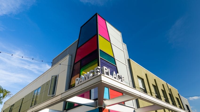 The corner of a building reading "Dance Place." The front of the corner has large square blocks of color while the rest of the building is tan with tall windows.