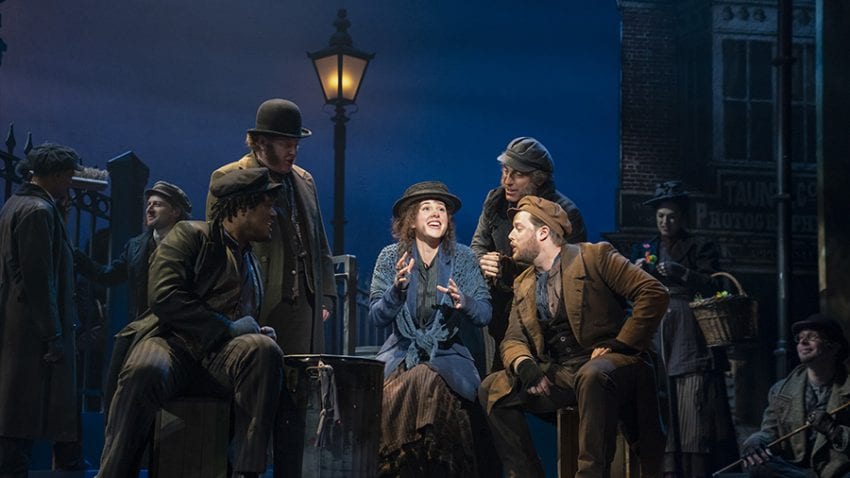 Actors in My Fair Lady perform a song. The actress playing Eliza sings in the center of several male actors with an excited expression. She wears a blue, ragged coat in front of a light post.