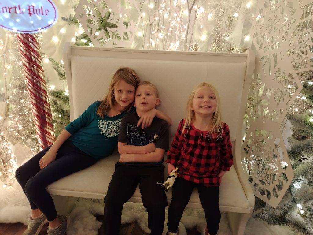 Three young kids sit on a white bench with sparkling white fake trees behind them. 