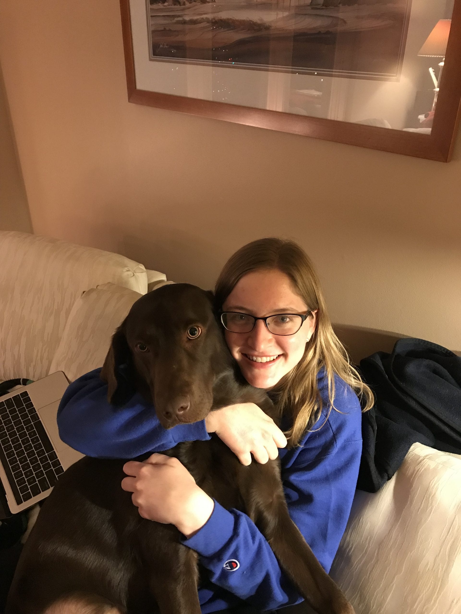 A chocolate lab sits on her owner, while the owner hugs her and smiles at the camera.