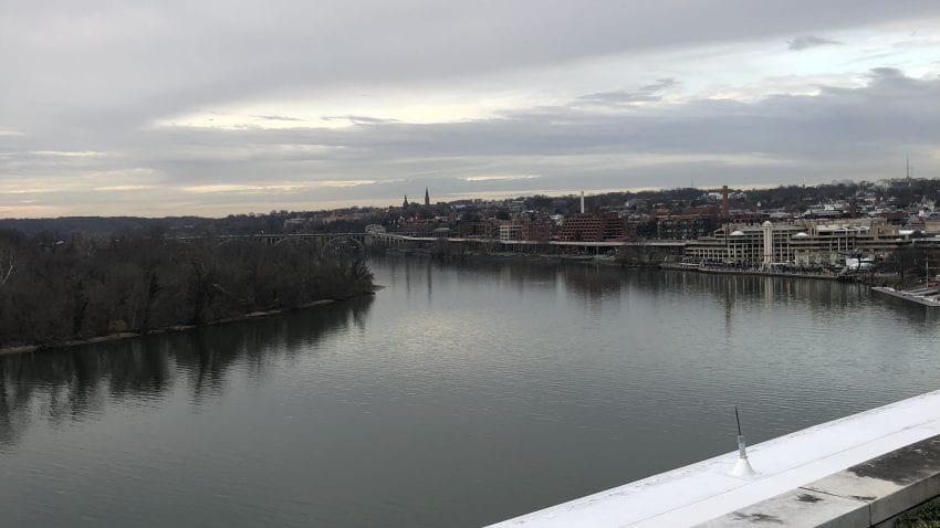 A view of a wide river from the top of the Kennedy Center. On the left side, there is a vast forest. On the right side, buildings from the neighborhood of Georgetown create a small skyline 