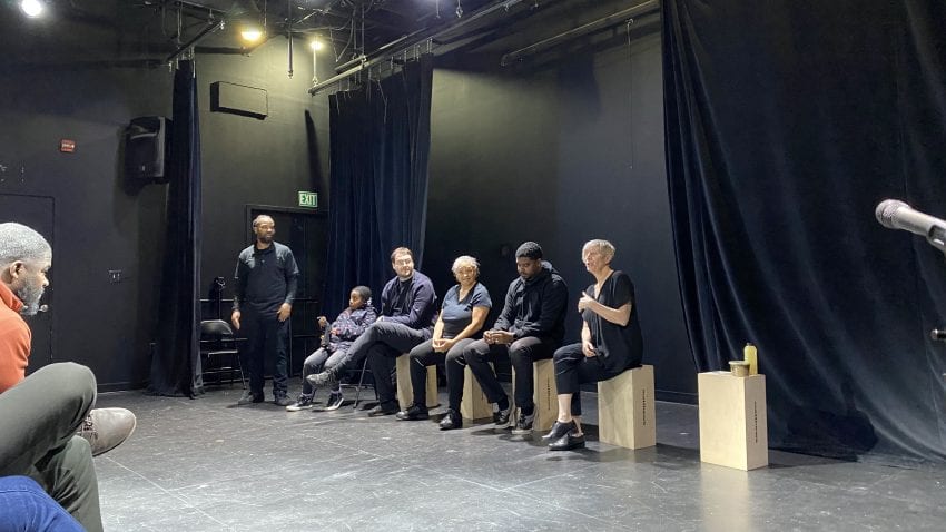 Six actors in black clothing sit on wooden boxes in a black-box style theater, with a black curtain in the back