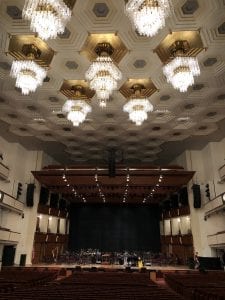 One of the concert halls in the Kennedy Center. Crystal lights hang from the ceiling and the stage features a large brown overlay. 