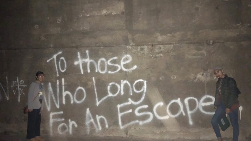 A gray wall with the words "to those who long for an escape" spray painted on the wall. Two students stand on either side of the text. 