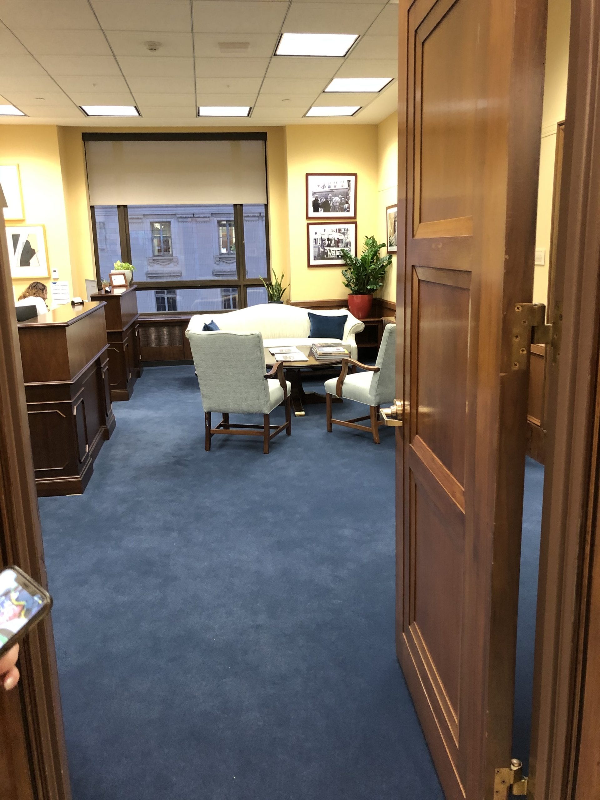 A brown door leading to an office with blue carpeting and a table with white chairs. A large desk sits on the left side.