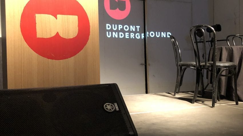 A stage with a podium with the red Dupont Underground symbol. A black table with black chairs sits on the right side. 