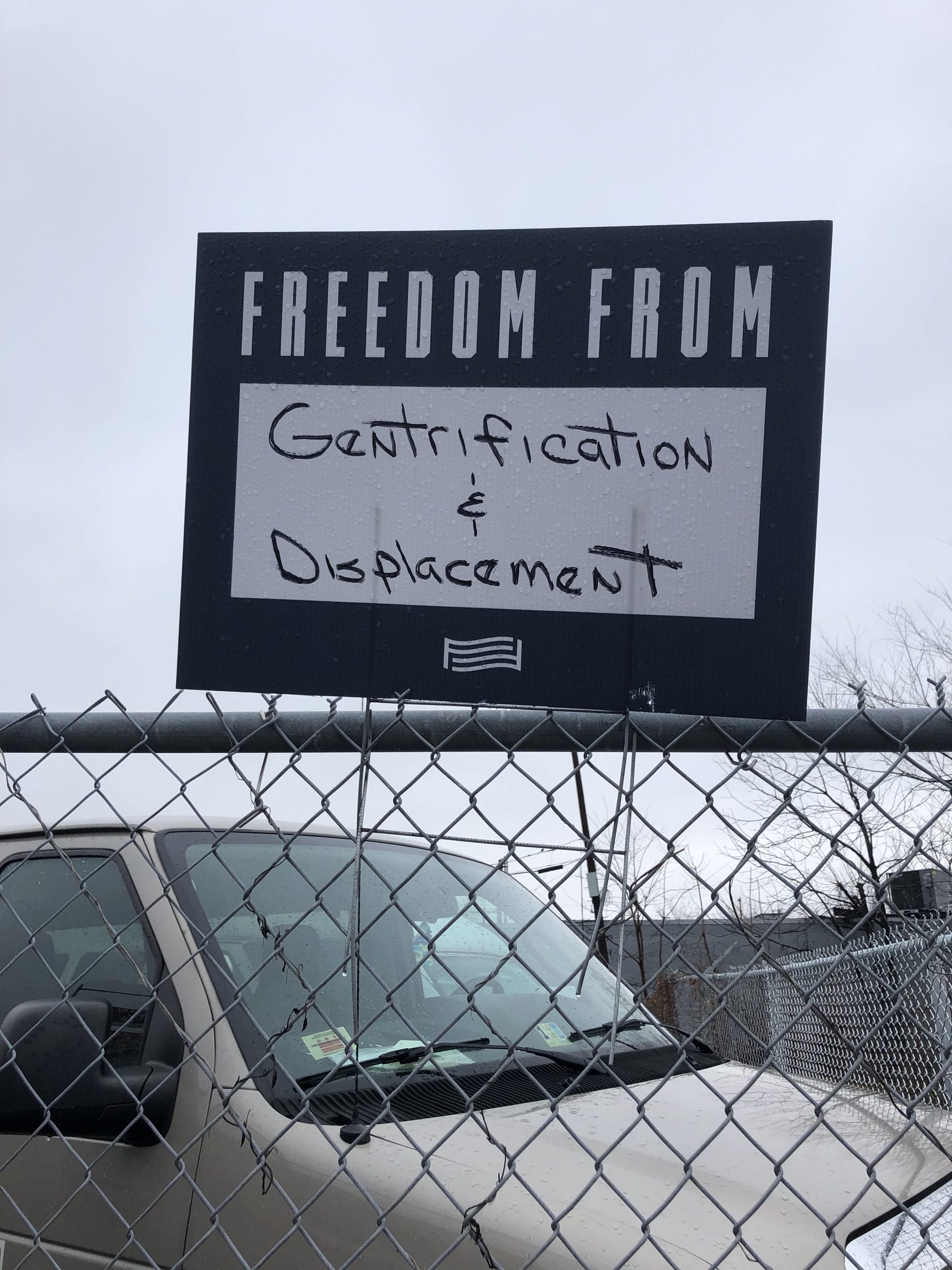 A sign reading "Freedom From: Gentrification and Displacement" on top of a car in front of a chain link fence