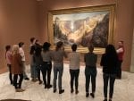 A group stands in front of a large painting. The painting is a framed image of a landscape.