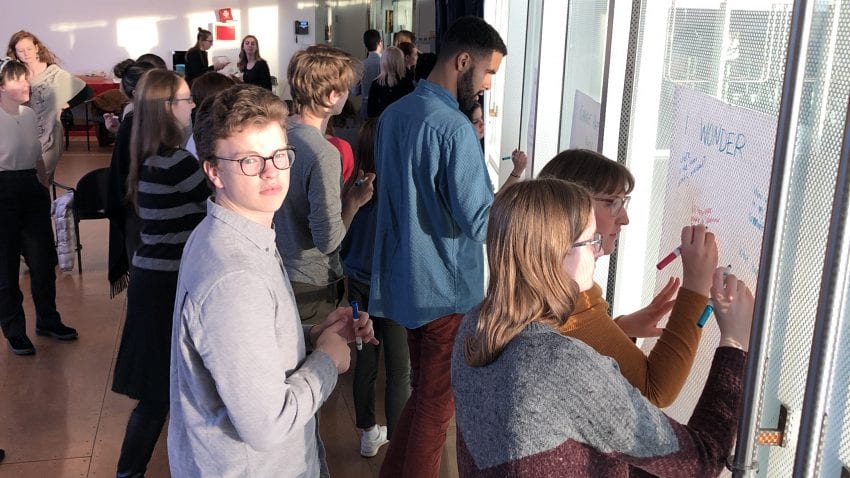 Students stand in a cluster and write on pieces of paper taped to glass windows. 