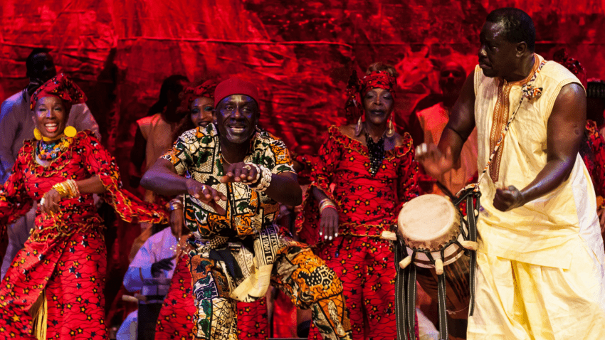 In front of a red background, dancers perform in colorful costumes and play on drums.