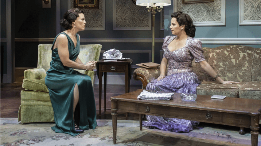 Two actresses in elegant dresses sit on living room seating. They are engrossed in conversation.