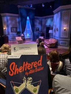 A photo of a program for the play "Sheltered" in front of the stage and audience. The stage features a living room with seating and an open window. The audience is full and sitting, ready for the performance. 