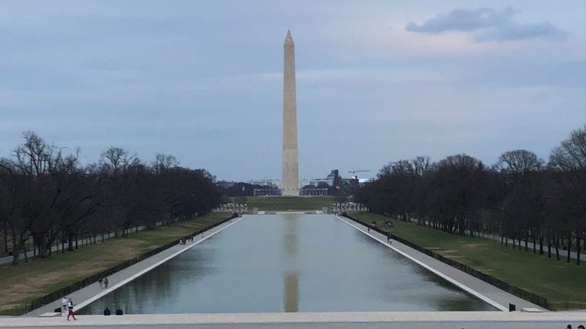 Washington Monument on National Mall in background, reflected in reflection pool. A few people walk around perimeter of pool. 