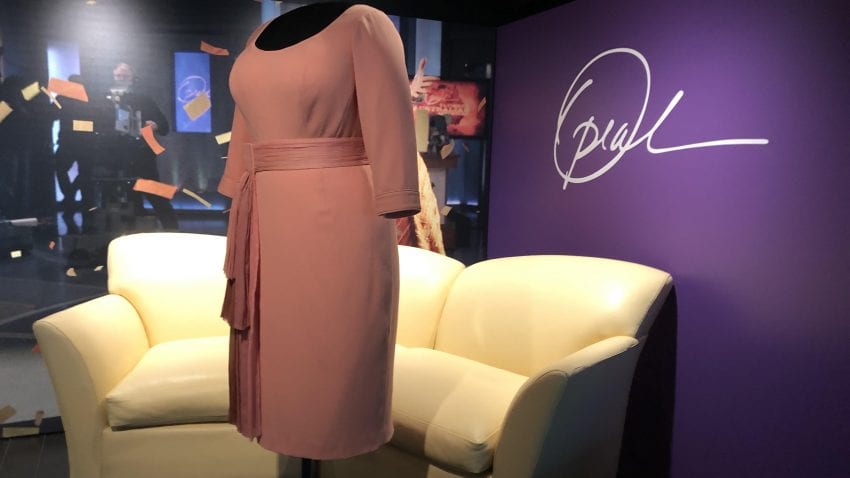 A light pink dress on display in front of a tan couch with a purple wall in the back with Oprah's signature on it