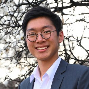 Portrait of Grayson Chan wearing a dress shirt and suit jacket standing in front of a flowering tree.