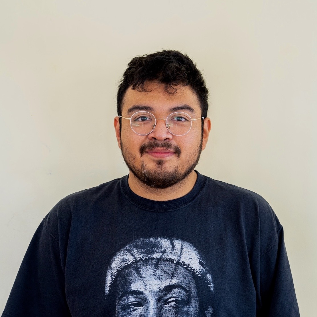 Latino man with beard and glasses standing in front of a wall.