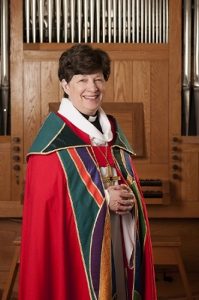 Bishop Elizabeth Eaton, wearing a traditional red robe, stands smiling at the camera.