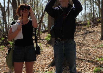 Two students look through binoculars in the woods.