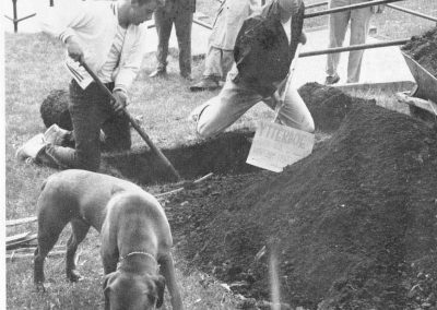 Two students dig a grave, with a few onlookers and another dog.